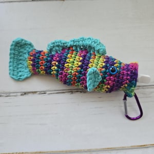 May include: A crocheted rainbow fish with a blue eye and a purple carabiner clip. The fish is made with a variety of colorful yarns.
