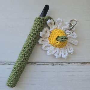 May include: A green crocheted pen holder and a white and yellow crocheted daisy keychain with a silver clasp. The pen holder is a solid green color and the daisy has a yellow center and white petals.