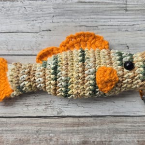 May include: A crocheted fish-shaped item with orange fins and tail, and a black eye. The body is a mix of beige, green, and brown yarn. A small orange metal clasp is attached to the fish.