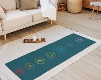 Puede incluir: Esterilla de yoga turquesa con siete símbolos de chakra en un degradado arcoíris. Los símbolos están dispuestos en línea sobre la esterilla. La esterilla es rectangular y está sobre una alfombra blanca.