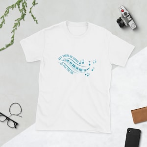 May include: White t-shirt with a musical theme. The design features a piano keyboard and musical notes in a teal color, accompanied by the text "Let there be songs to fill the air."