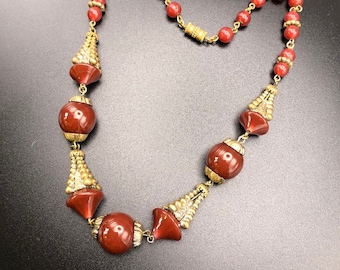 Op de afbeelding: Een rode en gouden kralenketting met grote, ronde, rode kralen en gouden accenten. De ketting heeft een kwastachtig ontwerp aan de uiteinden.
