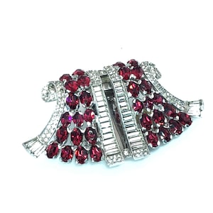 Könnte beinhalten: Eine Vintage-Brosche mit silberfarbenem Metallrahmen. Die Brosche hat einen Mittelteil aus klaren Kristallen im Baguetteschliff, flankiert von Reihen oval geschliffener, tiefroter Kristalle. Gebogene, bandartige Elemente sind mit klaren Kristallen besetzt.