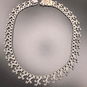 Op de afbeelding: Een zilveren ketting met een delicaat ontwerp met een herhalend patroon van kleine, vierkante stenen en ingewikkelde, draaiende details. De ketting is gemaakt van een ketting met een sluiting.