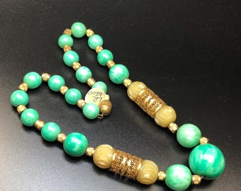 Könnte beinhalten: Eine grüne und goldene Perlenkette mit zwei großen, dekorativen goldenen und cremefarbenen Perlen. Die Kette hat ein Etikett mit der Aufschrift "LR DELCO".