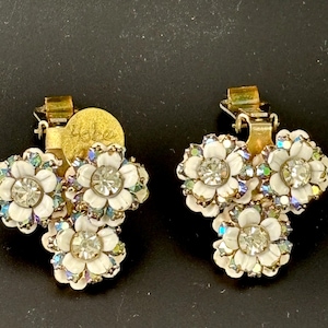 Puede incluir: Un par de pendientes de clip vintage Hobe con tres diseños de flores blancas con diamantes de imitación transparentes. Los pendientes son de color dorado y tienen un cierre de clip.