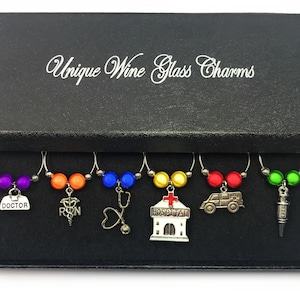 May include: Set of six wine glass charms in a black box. The charms feature different medical symbols, including a doctor's bag, a stethoscope, a hospital building, an ambulance, a syringe, and a caduceus. The charms are made of metal and have colorful beads.