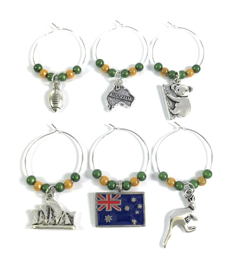 May include: Set of six Australian themed wine glass charms. Each charm features a different Australian icon, including a koala, kangaroo, rugby ball, the Sydney Opera House, the Australian flag, and a map of Australia. The charms are made of silver metal and have green and gold beads.