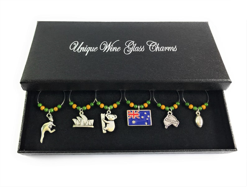 May include: Set of six wine glass charms in a black box. Each charm features a different Australian icon, including a kangaroo, the Sydney Opera House, a koala, the Australian flag, the outline of Australia, and a rugby ball. The charms are made of metal and have green and orange beads.