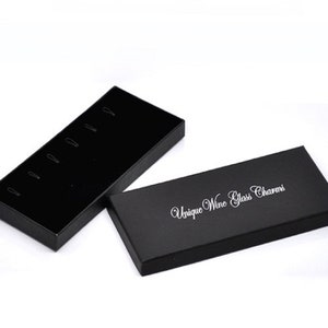May include: Black box with a black velvet interior and slots for holding wine glass charms. The box is open and the lid has the text "Unique Wine Glass Charms" printed on it.