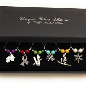 May include: Set of six wine glass charms in a black box. Each charm features a different winter-themed design, including mittens, a ribbon, skis, a snowflake, a skier, and a charm that says "Love to Ski".