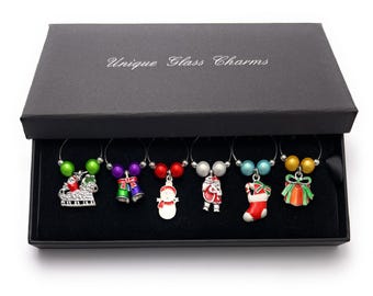 May include: A set of six Christmas-themed wine glass charms in a black box. The charms feature a snowman, Santa Claus, a stocking, a sleigh, bells, and a gift box. Each charm has a silver metal ring and two colorful beads.