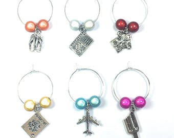 May include: Six silver wine glass charms with colorful beads and various travel-themed charms. The charms include flip flops, a postcard, a tropical shirt, a passport, an airplane, and an ice cream cone.