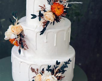 Op de afbeelding: Een witte taart met drie lagen en een druipglazuur. De taart is versierd met bloemstukken met oranje, crème en donkergroene accenten. De taart staat op een lichtgroen oppervlak tegen een donkergroene achtergrond.