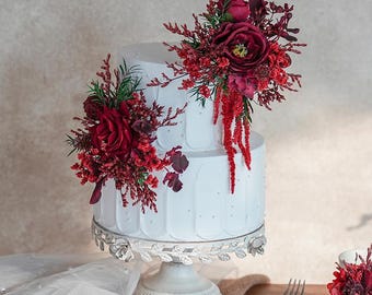 May include: A two-tiered white cake decorated with red flowers and greenery sits on a silver cake stand. A fork, knife, and small floral arrangement are on a wooden table. A cup and saucer are also on the table.