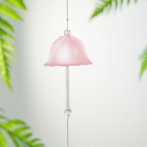 Pode incluir: Um delicado sino de vento rosa claro com design em forma de sino. Pendurado por um fio fino com contas transparentes. Folhas verdes de samambaia no fundo, criando uma est&eacute;tica suave e natural. Objeto decorativo.