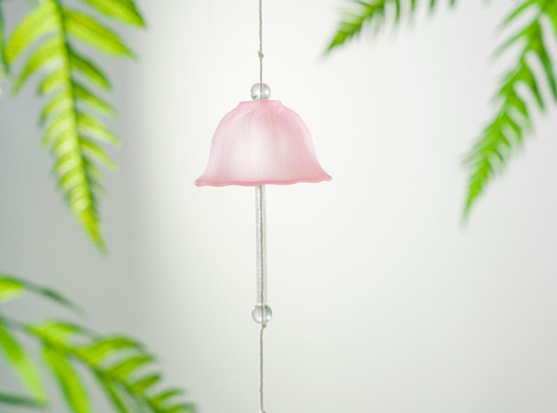 Pode incluir: Um delicado sino de vento rosa claro com design em forma de sino. Pendurado por um fio fino com contas transparentes. Folhas verdes de samambaia no fundo, criando uma est&eacute;tica suave e natural. Objeto decorativo.