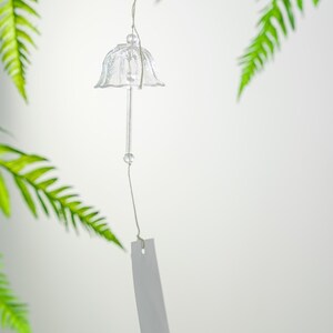 Pode incluir: Um sino de vento de vidro transparente com um topo em forma de sino e uma haste longa e fina. Uma pequena conta transparente &eacute; presa &agrave; haste. O sino pende de um fio fino, com uma etiqueta retangular branca na parte inferior. Folhas verdes de samambaia no fundo.
