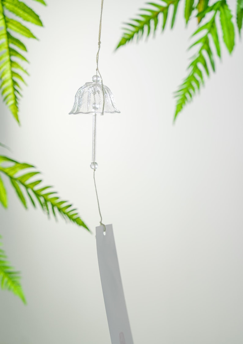 Pode incluir: Um sino de vento de vidro transparente com um topo em forma de sino e uma haste longa e fina. Uma pequena conta transparente &eacute; presa &agrave; haste. O sino pende de um fio fino, com uma etiqueta retangular branca na parte inferior. Folhas verdes de samambaia no fundo.