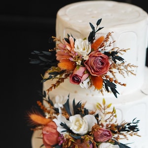 May include: A white wedding cake with three tiers decorated with orange and pink flowers, dried grasses, and dark green leaves.