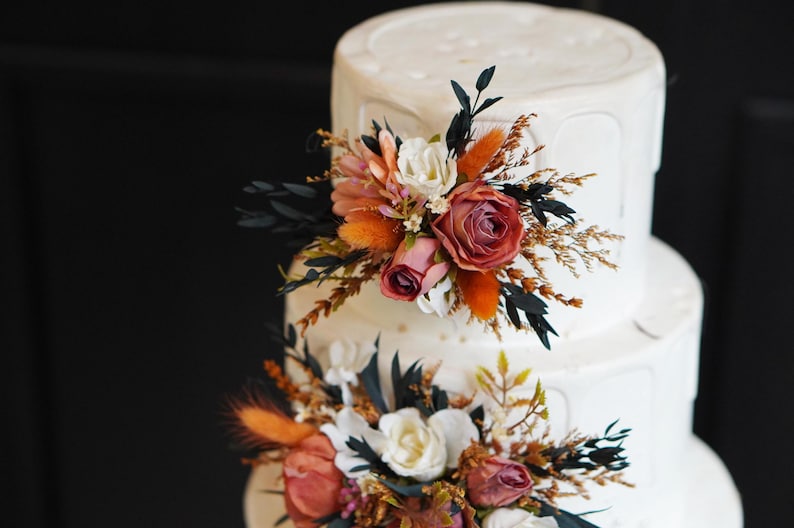 May include: A white wedding cake with three tiers decorated with orange and pink flowers, dried grasses, and dark green leaves.