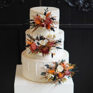 Può includere: Una torta nuziale bianca a tre piani decorata con fiori arancioni, rossi e bianchi. La torta è su un supporto per torte bianco.