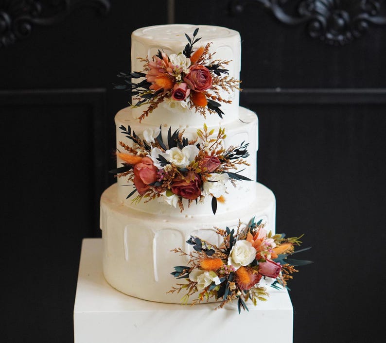 May include: A three-tiered white wedding cake decorated with orange, red, and white flowers. The cake is on a white cake stand.
