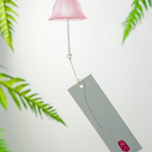 Pode incluir: Um delicado sino de vento rosa com um topo em forma de sino e uma etiqueta cinza. O sino de vento &eacute; suspenso por um fio fino, com folhas de samambaia verdes ao fundo. A etiqueta tem um selo vermelho.