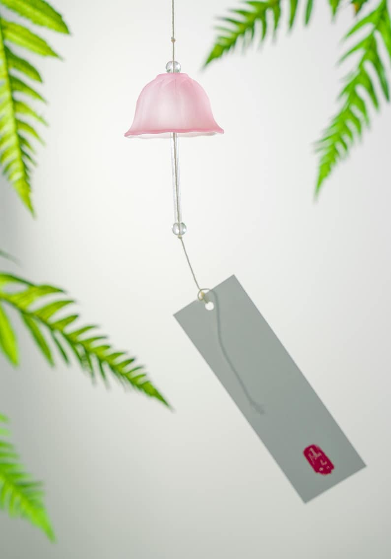 Pode incluir: Um delicado sino de vento rosa com um topo em forma de sino e uma etiqueta cinza. O sino de vento &eacute; suspenso por um fio fino, com folhas de samambaia verdes ao fundo. A etiqueta tem um selo vermelho.