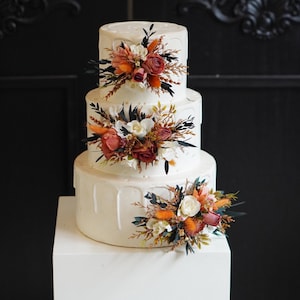 May include: A three-tiered white wedding cake decorated with dried flowers in shades of orange, red, and white. The cake is on a white cake stand.