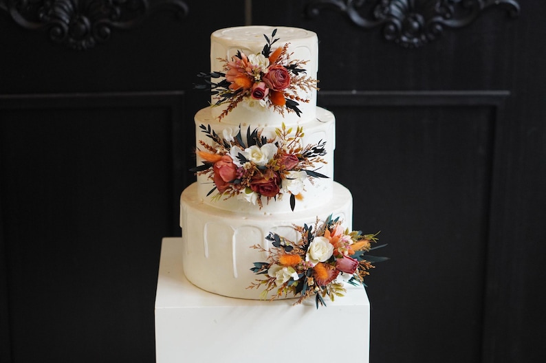May include: A three-tiered white wedding cake decorated with dried flowers in shades of orange, red, and white. The cake is on a white cake stand.