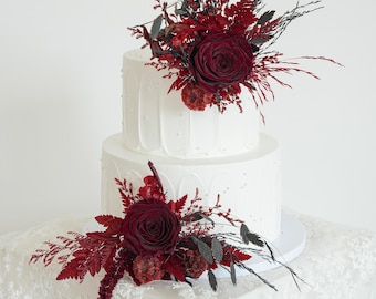 May include: A two-tiered white cake decorated with red roses and dark red and black foliage. The cake sits on a white lace tablecloth. The top tier is adorned with a large rose and floral arrangement.