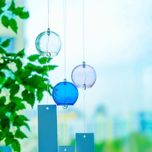 May include: Three spherical glass wind chimes in clear, blue, and light purple hanging from a wooden branch. Each glass sphere is suspended by a thin string attached to a rectangular white paper tag featuring a small red stamp at the bottom.