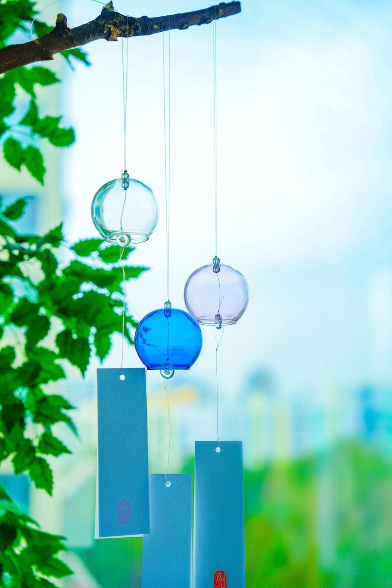 May include: Three spherical glass wind chimes in clear, blue, and light purple hanging from a wooden branch. Each glass sphere is suspended by a thin string attached to a rectangular white paper tag featuring a small red stamp at the bottom.