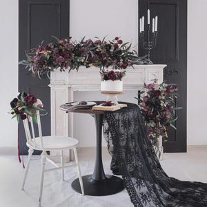 May include: A wedding reception scene featuring a white cake decorated with burgundy flowers, placed on a round table covered with a black lace tablecloth. A white chair with a bouquet and a floral arrangement on a mantelpiece.