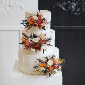 May include: A three-tiered white wedding cake decorated with dried flowers in shades of orange, red, and white. The cake is on a white cake stand.