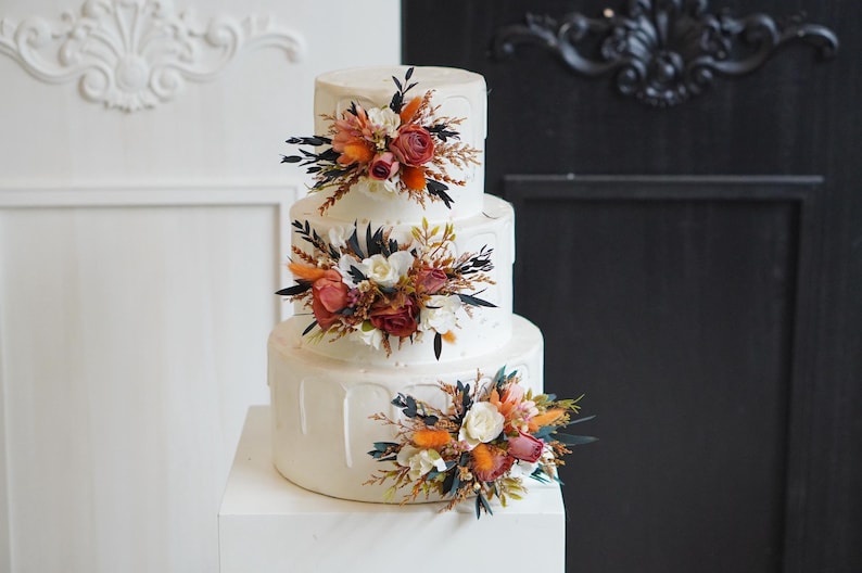 May include: A three-tiered white wedding cake decorated with dried flowers in shades of orange, red, and white. The cake is on a white cake stand.