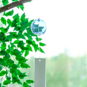 May include: A light blue spherical glass wind chime hanging from a branch. The chime features a clear glass clapper inside the sphere and a long, rectangular white paper strip attached by a thin string, marked with a small red Japanese character.