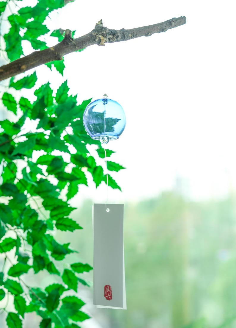 May include: A light blue spherical glass wind chime hanging from a branch. The chime features a clear glass clapper inside the sphere and a long, rectangular white paper strip attached by a thin string, marked with a small red Japanese character.