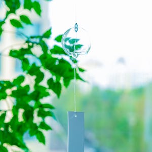 May include: A clear glass wind chime featuring a spherical bell suspended by a thin string. A rectangular white paper strip hangs from the bottom of the bell, displaying a small red circular stamp near the lower edge of the paper surface.