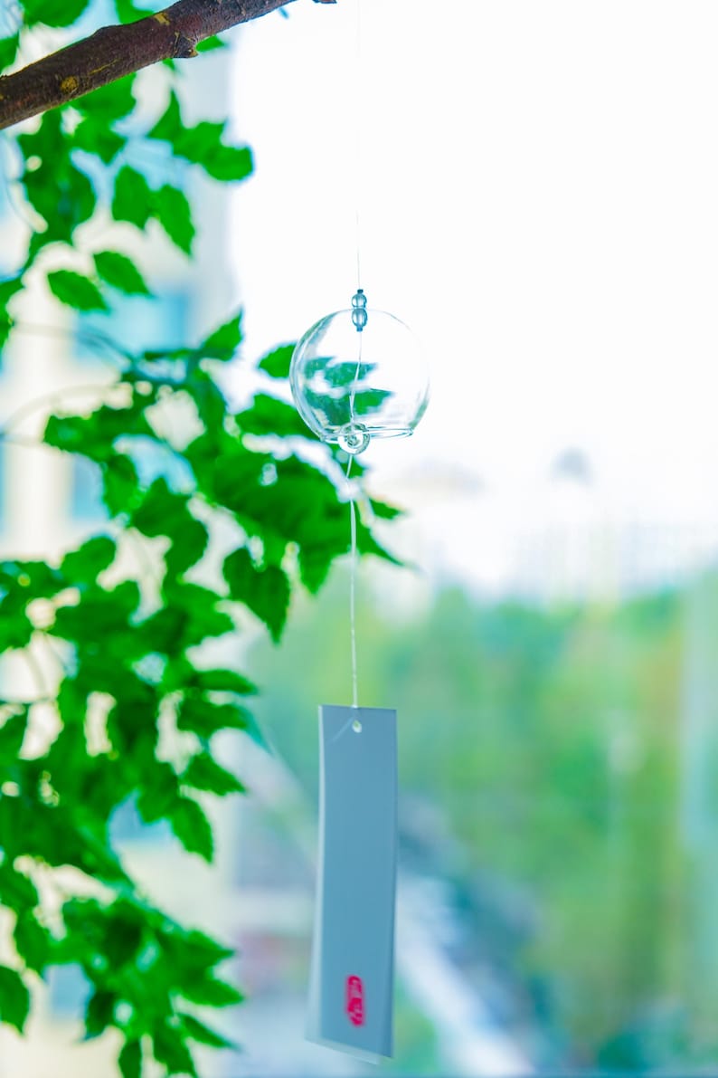 May include: A clear glass wind chime featuring a spherical bell suspended by a thin string. A rectangular white paper strip hangs from the bottom of the bell, displaying a small red circular stamp near the lower edge of the paper surface.