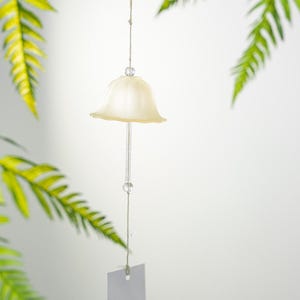 Pode incluir: Um sino de vento em forma de sino creme pendura-se de um fio fino, com duas contas de vidro transparente. O sino tem uma haste de vidro transparente e uma pequena etiqueta em branco na parte inferior. Folhas verdes de samambaia no fundo.
