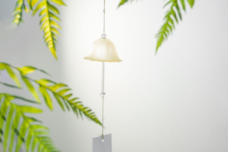 Pode incluir: Um sino de vento em forma de sino creme pendura-se de um fio fino, com duas contas de vidro transparente. O sino tem uma haste de vidro transparente e uma pequena etiqueta em branco na parte inferior. Folhas verdes de samambaia no fundo.
