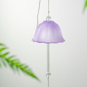 Pode incluir: Um delicado sino de vento cor de lavanda, com um topo em forma de sino e uma haste transparente pendurada. O sino tem uma superf&iacute;cie texturizada e &eacute; suspenso por um fio fino prateado. A folhagem verde est&aacute; desfocada no fundo.