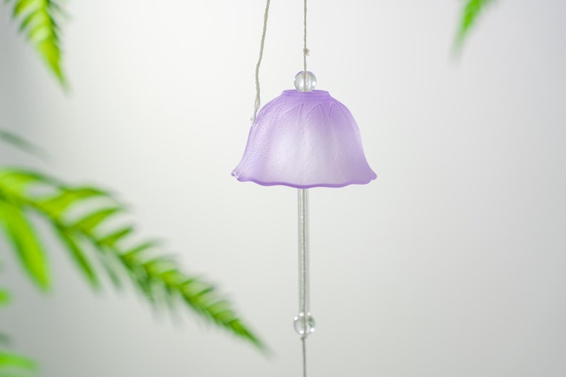 Pode incluir: Um delicado sino de vento cor de lavanda, com um topo em forma de sino e uma haste transparente pendurada. O sino tem uma superf&iacute;cie texturizada e &eacute; suspenso por um fio fino prateado. A folhagem verde est&aacute; desfocada no fundo.