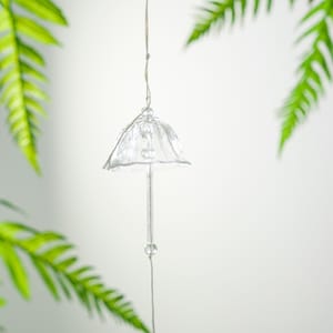 Pode incluir: Um sino de vento de vidro transparente, com um topo em forma de sino e uma haste longa e fina. O sino est&aacute; suspenso por um fio fino, com uma pequena conta esf&eacute;rica perto da base. Folhas verdes de samambaia emolduram o objeto.