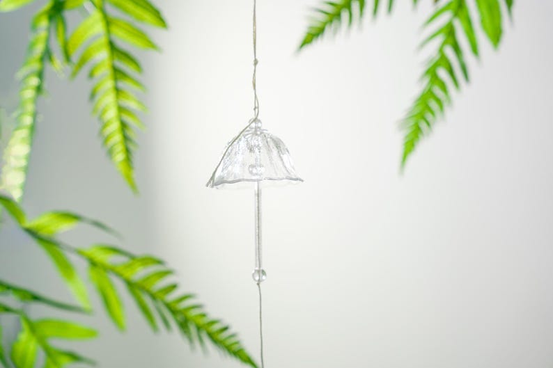 Pode incluir: Um sino de vento de vidro transparente, com um topo em forma de sino e uma haste longa e fina. O sino est&aacute; suspenso por um fio fino, com uma pequena conta esf&eacute;rica perto da base. Folhas verdes de samambaia emolduram o objeto.