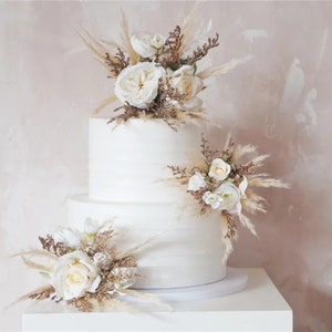 May include: Two-tier white wedding cake decorated with dried pampas grass and white roses.