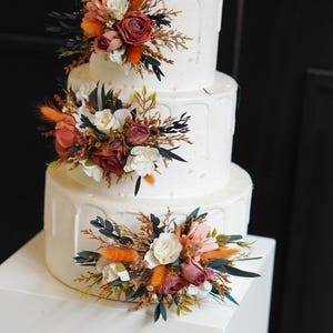 May include: A three-tiered white wedding cake decorated with cascading white frosting and floral arrangements. The flowers are a mix of white, orange, and pink.