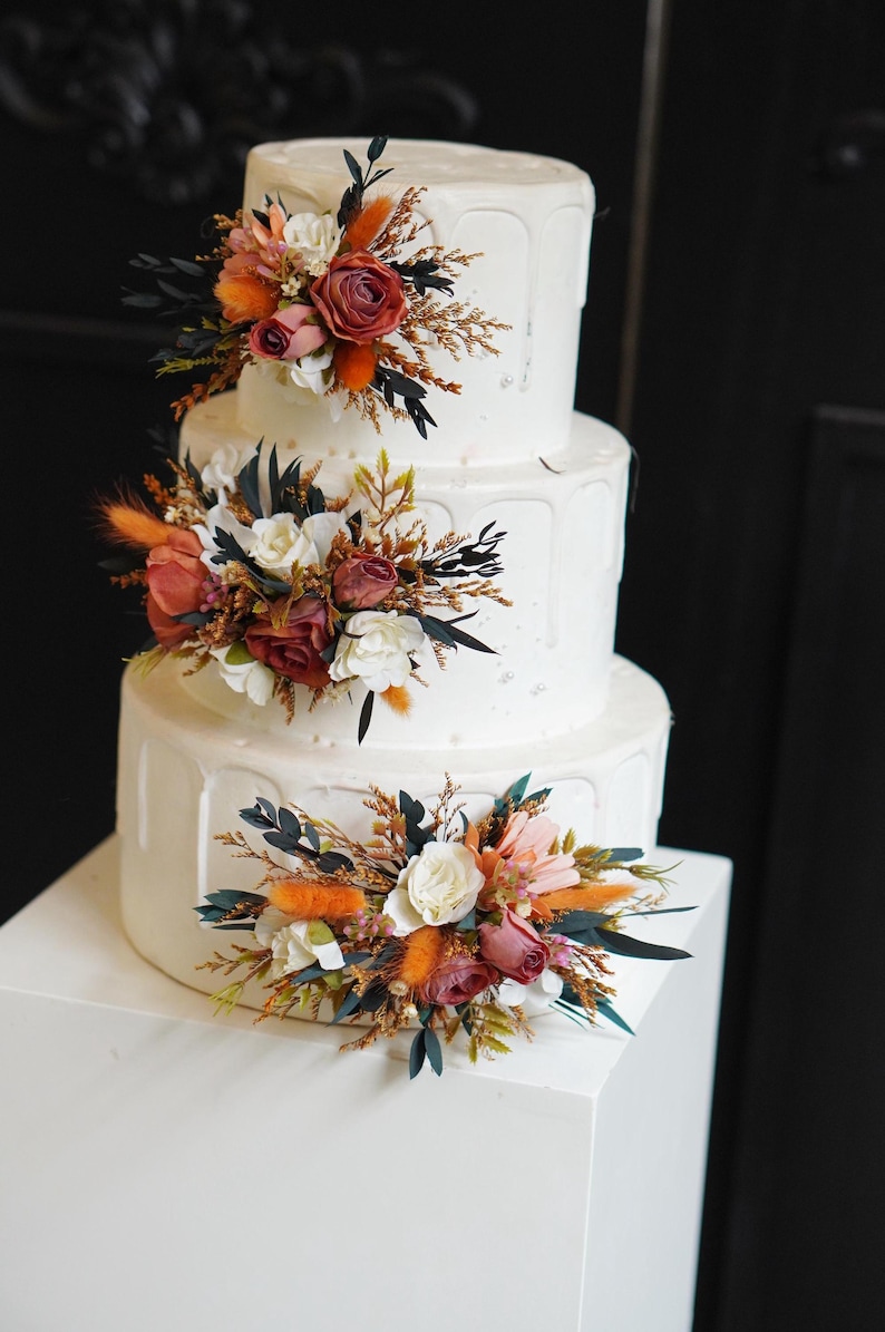 May include: A three-tiered white wedding cake decorated with cascading white frosting and floral arrangements. The flowers are a mix of white, orange, and pink.